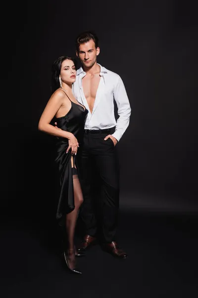 Homme en chemise déboutonnée debout avec la main dans la poche près de la femme en robe et bas sur noir — Photo de stock