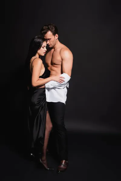 Full length view of elegant woman in satin dress taking white shirt off sexy man on black — Stock Photo