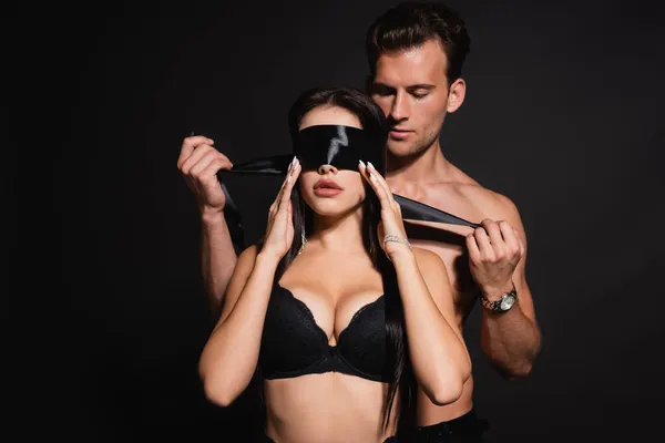 Sexy hombre atando seda ojos vendados en mujer en sujetador aislado en negro - foto de stock