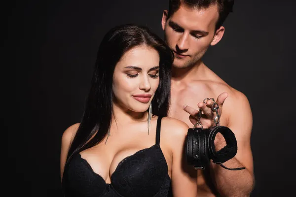 Young shirtless man holding handcuffs near smiling woman in bra isolated on black — Stock Photo