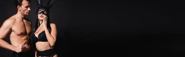 Sexy uomo toccare faccia di donna in maschera coniglietto con frusta isolato su nero, banner — Foto stock