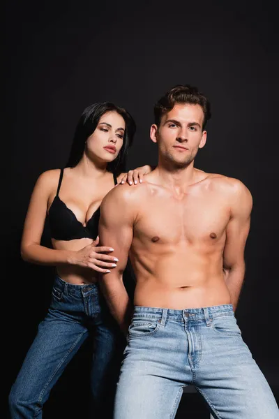 Sexy sin camisa hombre en jeans mirando cámara cerca sensual morena mujer aislado en negro - foto de stock