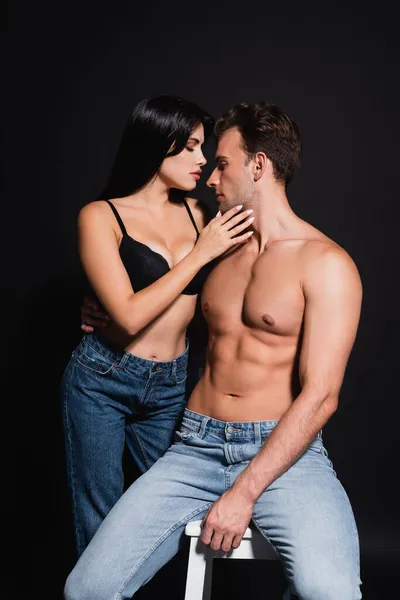 Sexy brunette woman seducing man with muscular torso sitting isolated on black — Stock Photo