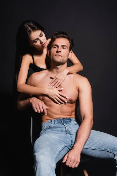 Страстная женщина в лифчике обнимает мускулистого мужчину сидящего изолированного на черном — стоковое фото
