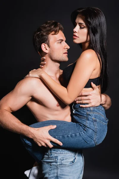 Vista lateral del hombre musculoso sin camisa sosteniendo a la mujer en sujetador y jeans aislados en negro — Stock Photo