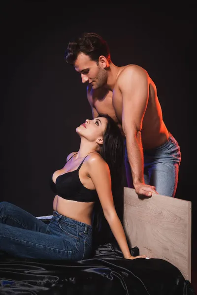 Sexy homme torse nu regardant femme séduisante assise sur la literie de soie en soutien-gorge et jeans isolés sur noir — Photo de stock