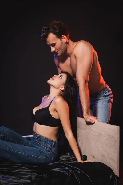 Sexy shirtless man looking at seductive woman sitting on silk bedding in bra and jeans isolated on black — стоковое фото