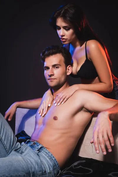 Sexy shirtless man sitting on bed near passionate woman seducing him isolated on black — Stock Photo