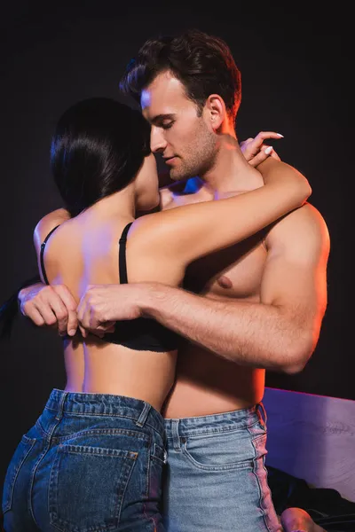 Young man unfastening bra of brunette woman in lighting isolated on black — Stock Photo