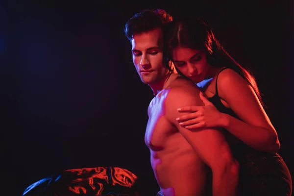 Sexy brunette woman hugging shirtless muscular man in red lighting isolated on black — Stock Photo