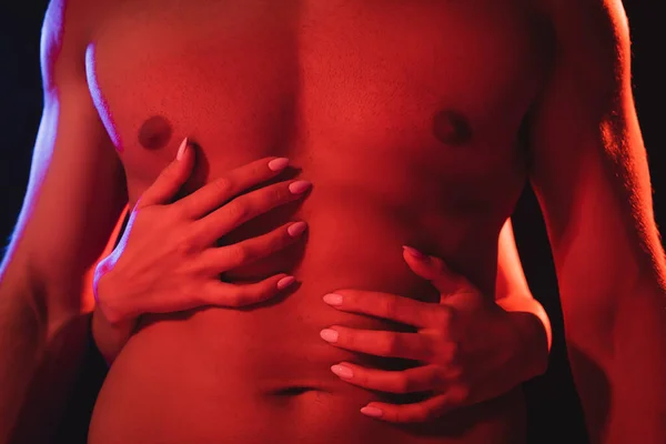 Vista recortada de la mujer abrazando el torso muscular del joven en la iluminación roja aislado en negro - foto de stock