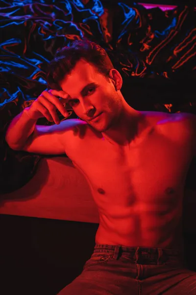 Sexy shirtless man looking at camera near black bedding in red lighting — Stock Photo