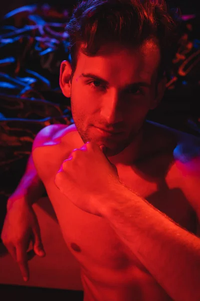 Young shirtless man looking at camera while posing near black blurred bedding in red lighting — Stock Photo