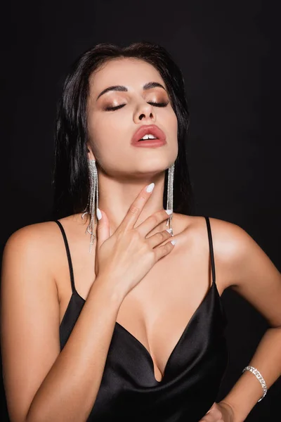 Sexy woman in silk dress and earrings touching neck while posing with closed eyes isolated on black — Stock Photo