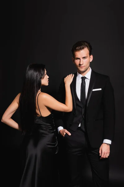 Brunette woman in silk dress touching shoulder of elegant man standing with hand in pocket on black — Stock Photo