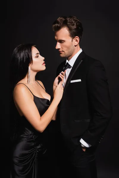 Brunette woman seducing man in elegant suit and touching his tie isolated on black — Stock Photo