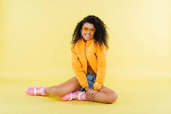 Full length of happy african american young woman in pink roller skates sitting on yellow — Stock Photo