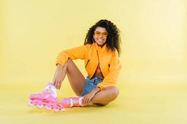 Longitud completa de alegre afroamericana joven mujer en rosa patines sentado en amarillo - foto de stock