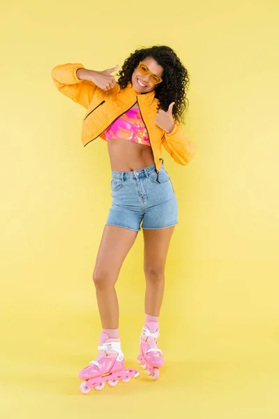 Comprimento total de alegre Africano americano jovem mulher em patins de rolo rosa mostrando polegares para cima no amarelo — Fotografia de Stock