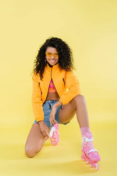 Comprimento total de sorrir Africano americano jovem mulher em patins de rolo rosa sentado no joelho em amarelo — Fotografia de Stock