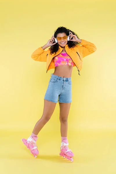 Pleine longueur de sourire afro-américaine jeune femme en patins à roulettes roses écouter de la musique sur jaune — Photo de stock