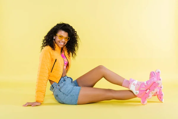 Pleine longueur de heureuse jeune femme afro-américaine en lunettes de soleil et patins à roulettes roses assis sur jaune — Photo de stock