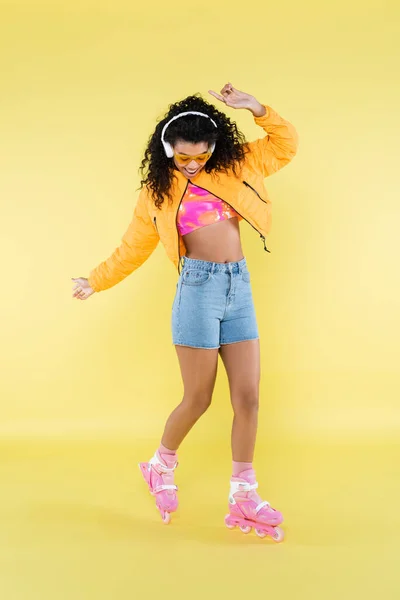 Full length of african american young woman in headphones and pink roller skates on yellow — Stock Photo