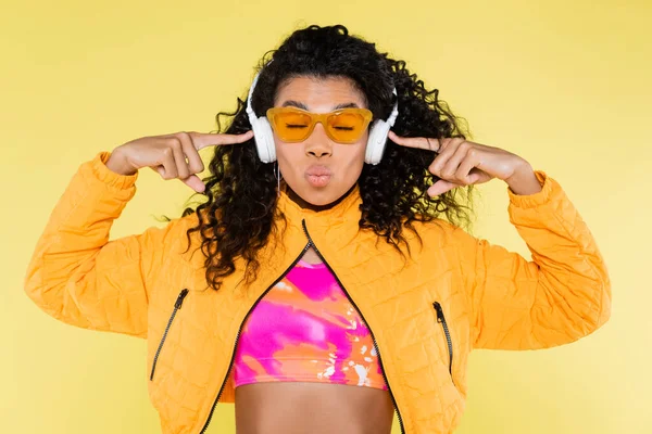 Curly african american young woman in sunglasses pointing at headphones and pouting lips isolated on yellow — Stock Photo