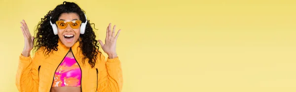 Espantada afro-americana jovem mulher em óculos de sol e fones de ouvido isolados em amarelo, banner — Fotografia de Stock