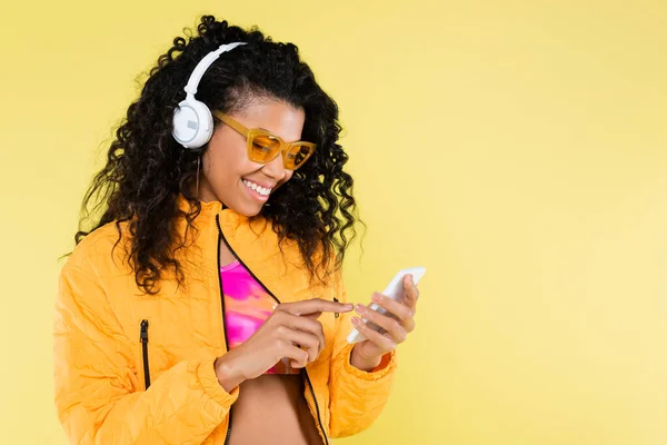 Feliz afro-americana jovem mulher em fones de ouvido usando smartphone isolado no amarelo — Fotografia de Stock