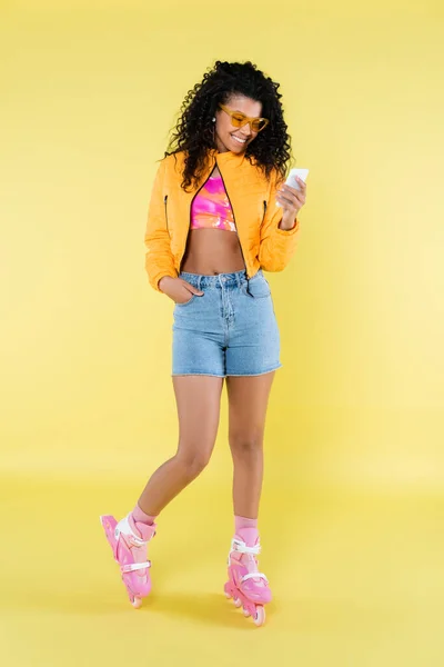 Longitud completa de mujer joven afroamericana en patines de color rosa posando mientras mira el teléfono inteligente en amarillo - foto de stock