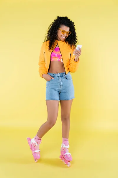 Full length of african american young woman in pink roller skates posing while looking at smartphone on yellow — Stock Photo