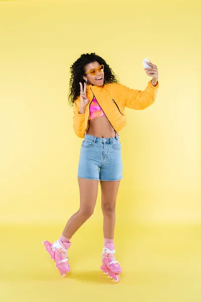 Pleine longueur de jeune femme afro-américaine étonnée sur patins à roulettes roses montrant le signe de la pièce tout en prenant selfie sur jaune — Photo de stock