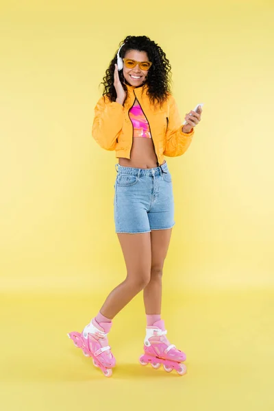 Comprimento total da jovem americana africana em patins de rolo rosa e fones de ouvido usando smartphone no amarelo — Fotografia de Stock
