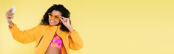 Happy african american young woman adjusting sunglasses while taking selfie isolated on yellow, banner — Stock Photo