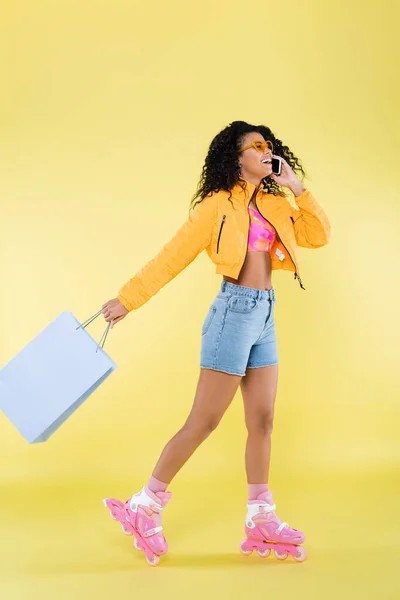 Afrikanisch-amerikanische junge Frau auf rosa Rollschuhen unterhält sich auf Handy, während sie Einkaufstasche auf gelb hält — Stockfoto