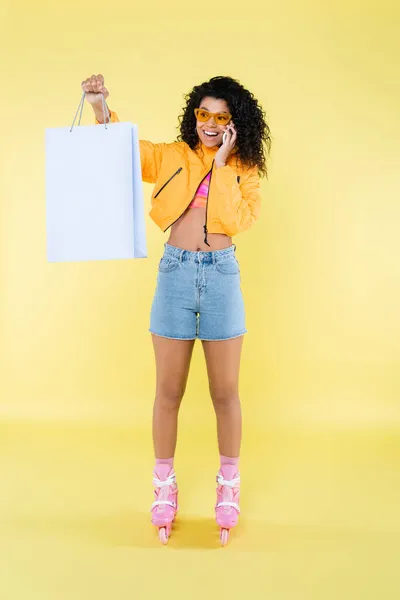 Piena lunghezza di felice afroamericana giovane donna sui pattini a rotelle parlando sul cellulare mentre tiene la borsa della spesa sul giallo — Foto stock