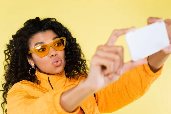 Rizado africano americano joven mujer en gafas de sol pucheros labios y tomando selfie aislado en amarillo - foto de stock