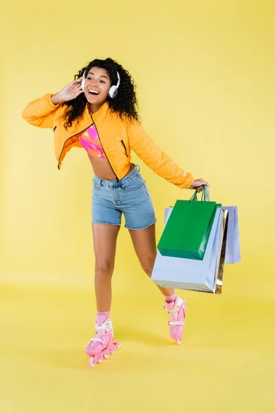 Повна довжина здивованої афроамериканської молодої жінки в роликових ковзанах і навушниках, що тримають сумки для покупок на жовтому — стокове фото