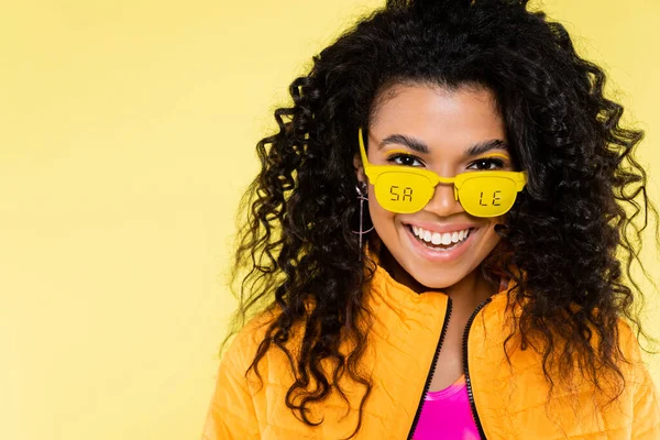 Feliz afro-americana jovem mulher em óculos de sol com venda lettering isolado no amarelo — Fotografia de Stock