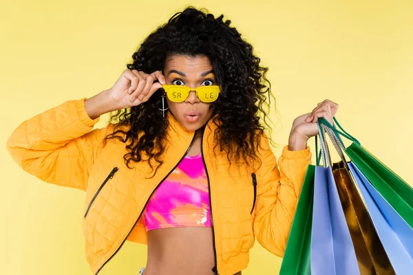 Sorpreso afroamericano giovane donna in occhiali da sole con la vendita lettering tenendo borse della spesa isolate su giallo — Foto stock
