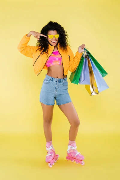 Pleine longueur de heureuse jeune femme afro-américaine sur patins à roulettes roses pointant vers les lunettes de soleil avec lettrage de vente sur jaune — Photo de stock
