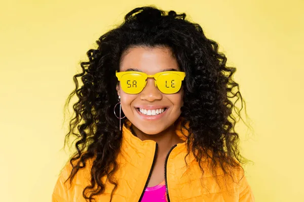 Sorridente afro-americano jovem mulher em óculos de sol com venda lettering isolado no amarelo — Fotografia de Stock