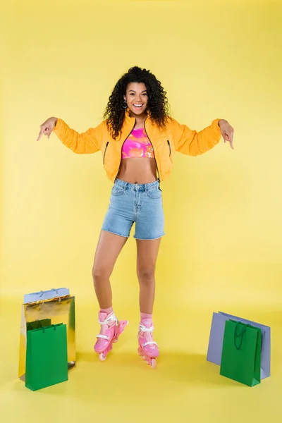 Longitud completa de la joven afroamericana feliz en patines de color rosa apuntando a las bolsas de compras en amarillo - foto de stock