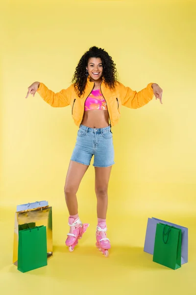 Pleine longueur de heureuse jeune femme afro-américaine en patins à roulettes roses pointant vers des sacs à provisions sur jaune — Photo de stock