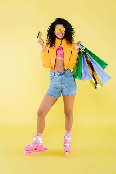 Longitud completa de la feliz mujer afroamericana en gafas de sol y patines con bolsas de compras y tarjeta de crédito en amarillo - foto de stock