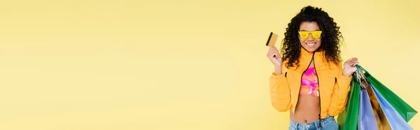 Mujer afroamericana alegre en gafas de sol con letras de la venta sosteniendo bolsas de compras y tarjeta de crédito aislada en amarillo, bandera - foto de stock