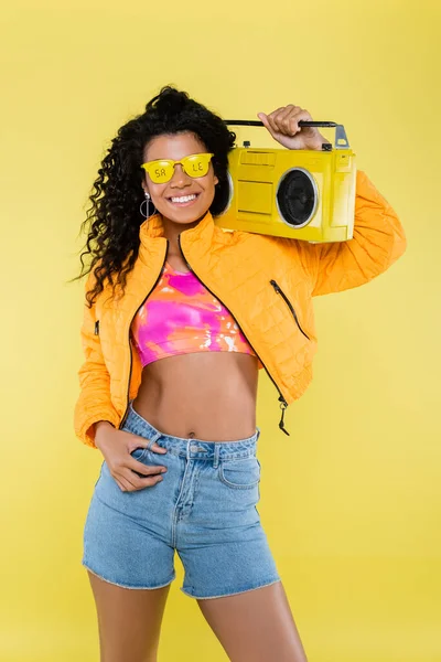 Sorridente Africano americano jovem mulher em óculos de sol com venda lettering segurando boombox isolado no amarelo — Fotografia de Stock