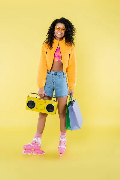 Full length of happy african american woman in sunglasses and roller skates holding shopping bags and boombox on yellow — Stock Photo