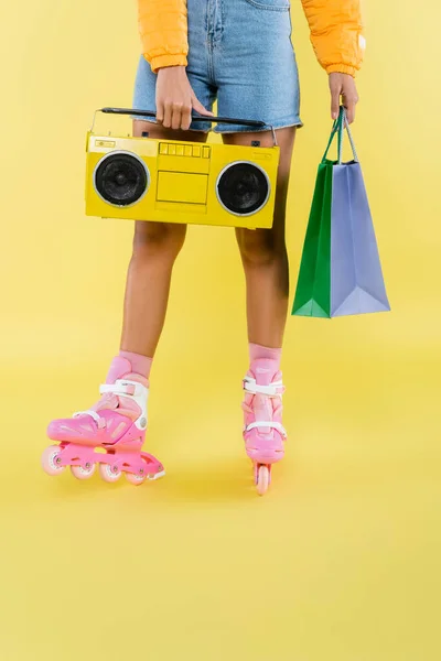 Vista recortada de mujer afroamericana en patines con bolsas de compras y boombox en amarillo - foto de stock